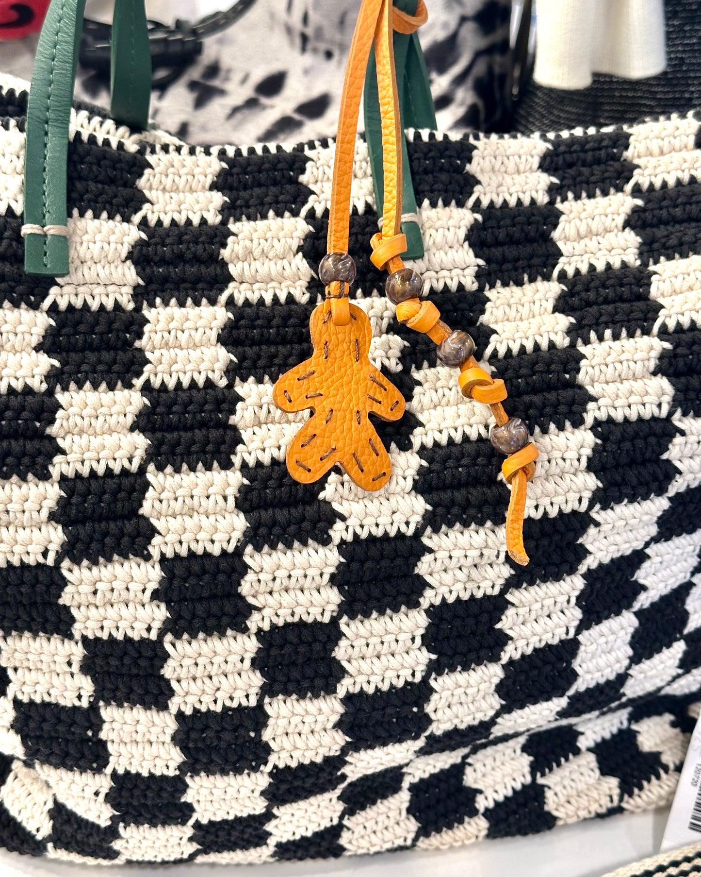 The Henry Beguelin Little Men Bag Charms feature a black and white checkered woven bag with green handles, yellow beads on one handle, an orange Italian leather gingerbread man-shaped charm, and a handmade logo.