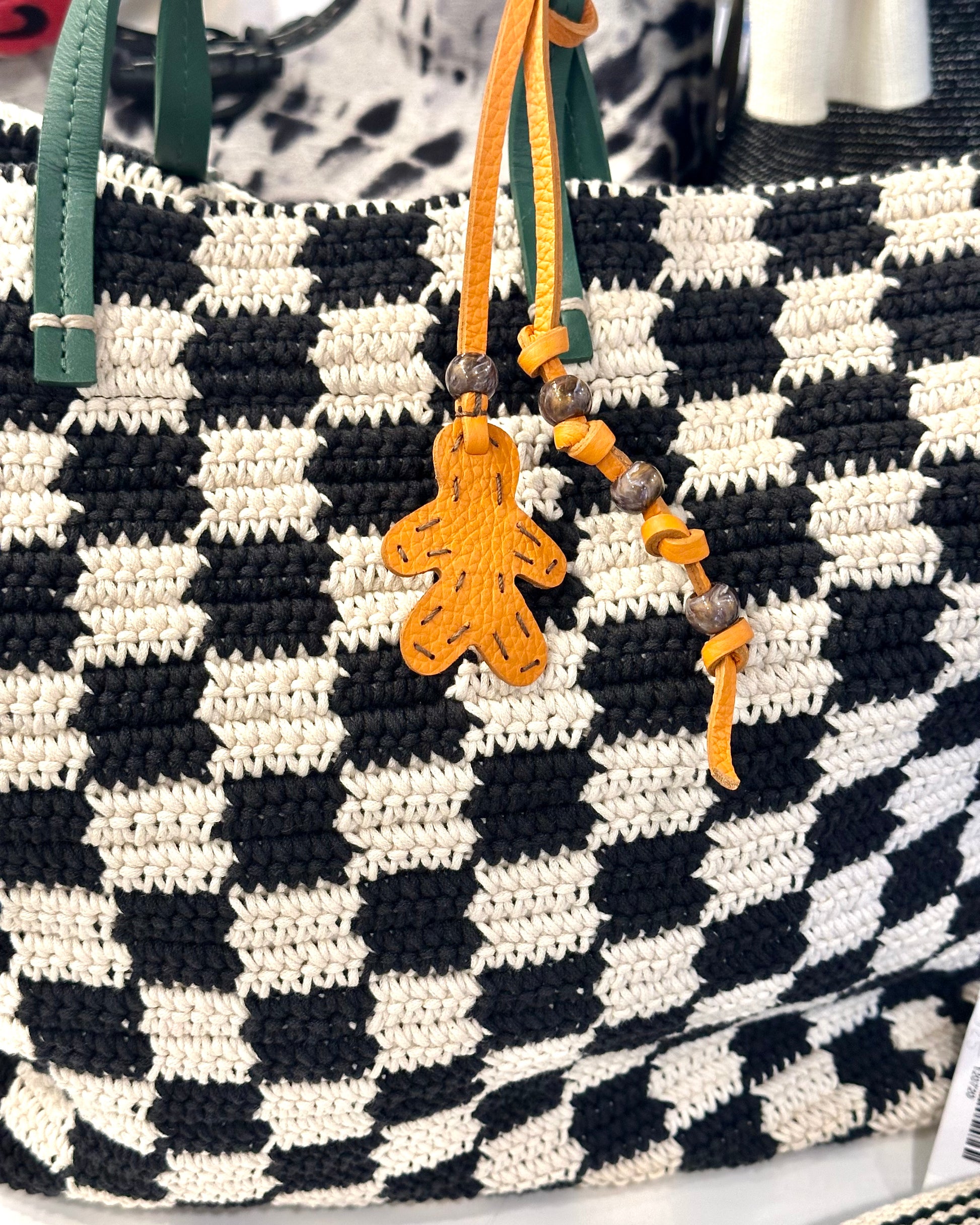 The Henry Beguelin Little Men Bag Charms feature a black and white checkered woven bag with green handles, yellow beads on one handle, an orange Italian leather gingerbread man-shaped charm, and a handmade logo.