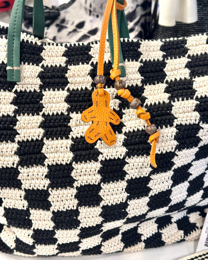 The Henry Beguelin Little Men Bag Charms feature a black and white checkered woven bag with green handles, yellow beads on one handle, an orange Italian leather gingerbread man-shaped charm, and a handmade logo.