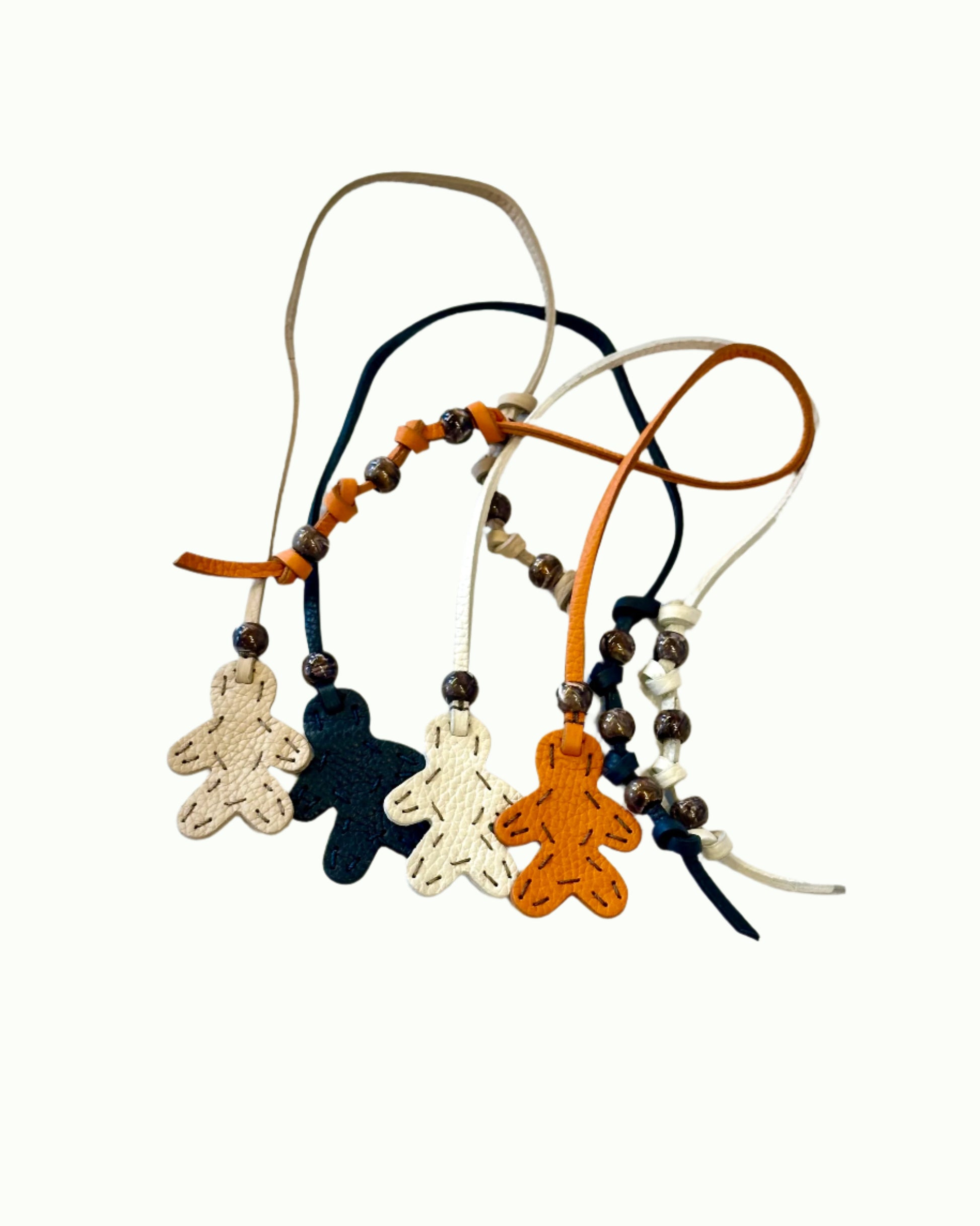 Four Little Men Bag Charms by Henry Beguelin—gingerbread-shaped figures in cream, dark blue, white, and orange Italian leather with beaded cords—are displayed on a white surface.