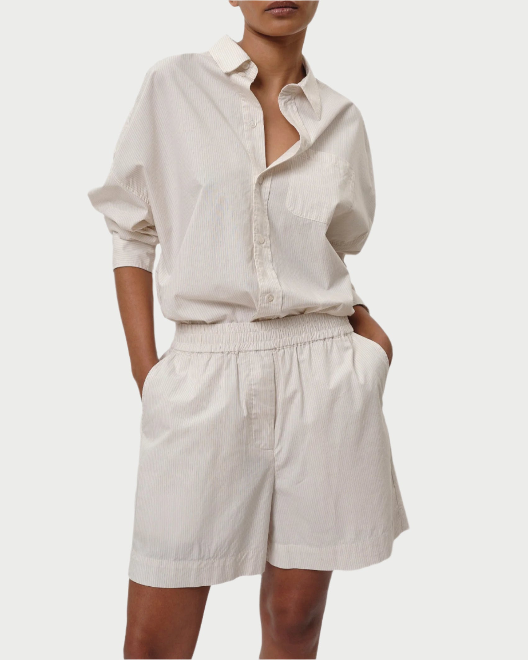 Sporting the Alma Shorts by Herskind, a person in a light beige, collared, long-sleeve shirt tucked in effortlessly pairs their look with high-waisted, relaxed-fit shorts. The 100% cotton shirt is slightly oversized and perfectly complements the sophisticated style as hands rest casually in pockets.