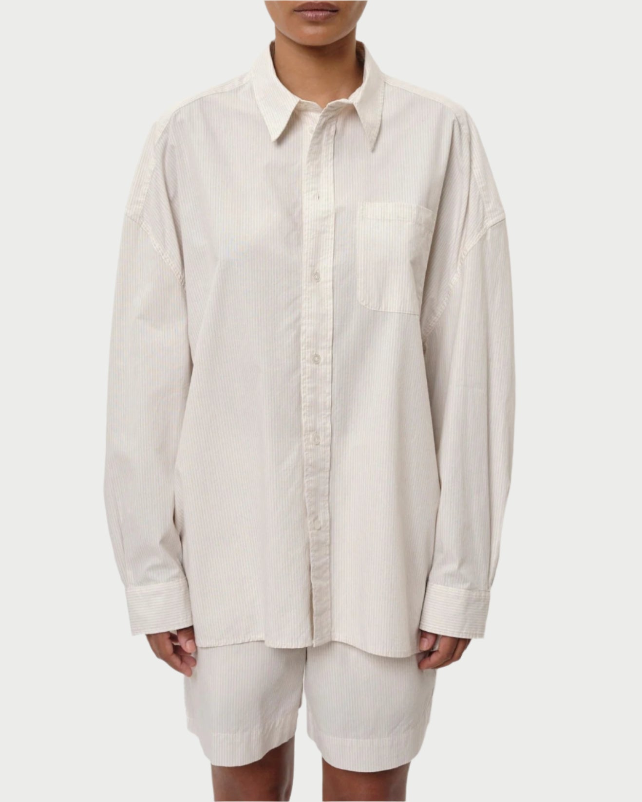 Someone sports a Herskind Lex Shirt, oversized and light beige with subtle vertical stripes and a front pocket. Its untucked long sleeves add effortless style, while perfectly paired with matching beige pants for an ideal Spring/Summer look.