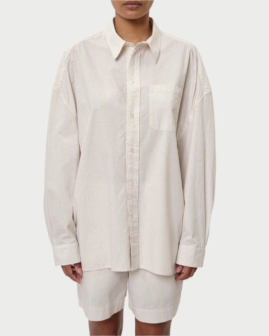 Someone sports a Herskind Lex Shirt, oversized and light beige with subtle vertical stripes and a front pocket. Its untucked long sleeves add effortless style, while perfectly paired with matching beige pants for an ideal Spring/Summer look.