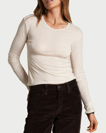 Woman wearing a white long-sleeve top and dark pants against a plain wall.