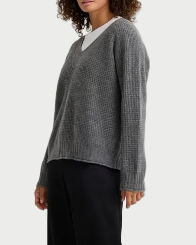 A person wearing a grey ribbed knit One Grey Day Linden Cashmere V-Neck over a white crewneck shirt and black trousers.