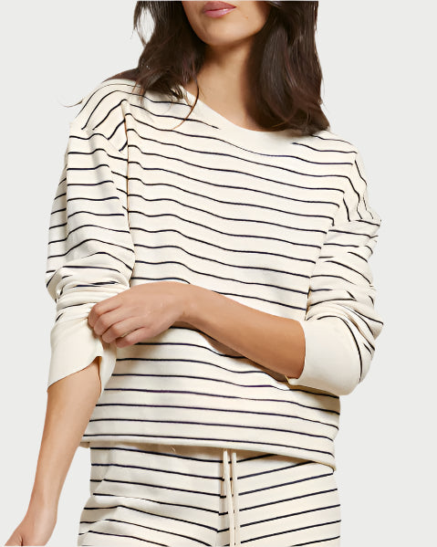 A woman wears the cream Tyler Terry Pullover by perfectwhitetee, a vintage French terry sweatshirt with thin black stripes and matching drawstring pants, standing with one arm crossed and facing slightly to the side.