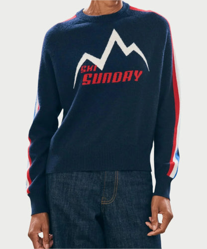 Someone is wearing the JUMPER Ski Sunday Sweater, a navy blue ski sweater showcasing a white and red mountain design with "SKI SUNDAY" emblazoned on the front. This cozy wool item features sleeves decorated with red and white stripes, complemented by dark denim jeans.