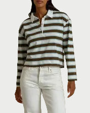A woman stands against a draped beige fabric backdrop wearing a long-sleeve knit polo-style top with dark olive and white horizontal stripes, white pants, and taupe shoes.