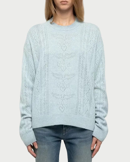 A woman stands against a light gray studio background wearing a pale icy blue cable-knit sweater, blue jeans, and white sneakers.