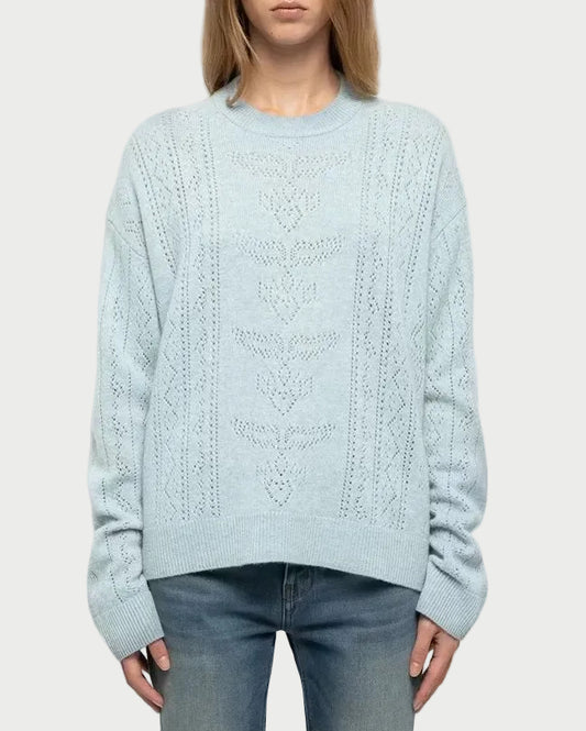 A woman stands against a light gray studio background wearing a pale icy blue cable-knit sweater, blue jeans, and white sneakers.