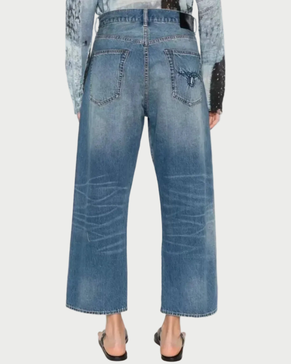 Back view of a model wearing high-waisted, wide-leg cropped blue denim jeans with a faded wash and a western-style embroidered patch on the right back pocket.