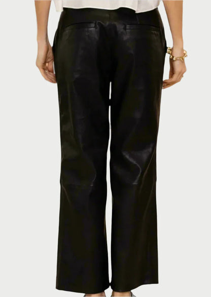 A person is wearing SPRWMN's black Cropped Baggy Leather Trousers and a white top, shown from the back. The shiny trousers feature a low-rise fit and fall just above the ankles. One hand rests by their side while the other showcases a gold bracelet.