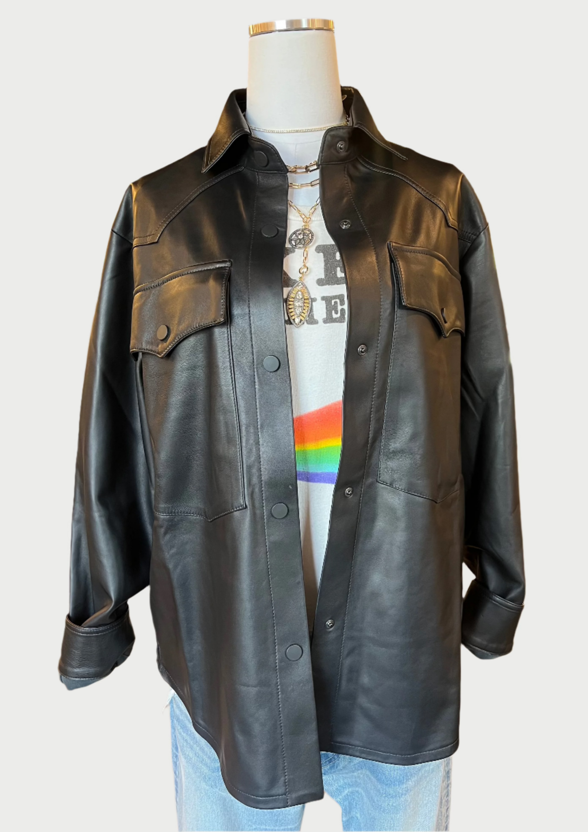 A mannequin dressed in a vintage black 3x1 Denim Leather Shirt Jacket over a white T-shirt with a rainbow stripe and gold layered necklaces. The outfit is completed with light-wash jeans. The background displays a clothing store with various garments and accessories.