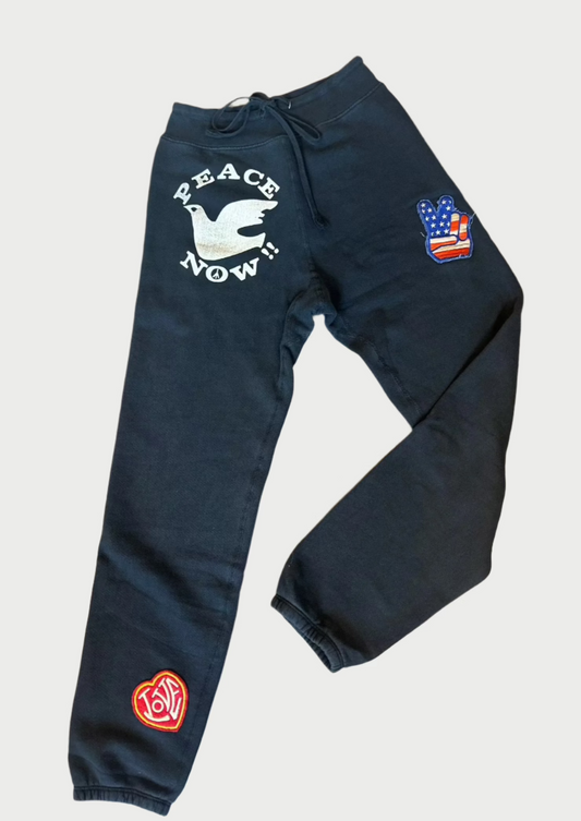 A pair of handmade Madeworn Peace Now Sweatpants is displayed on a wooden floor. The left leg features a white dove and the text "Peace Now!" on the thigh, and a circular red logo near the ankle. The right leg has a patch with an American flag and a peace sign on the thigh, all in a vintage unisex fit.