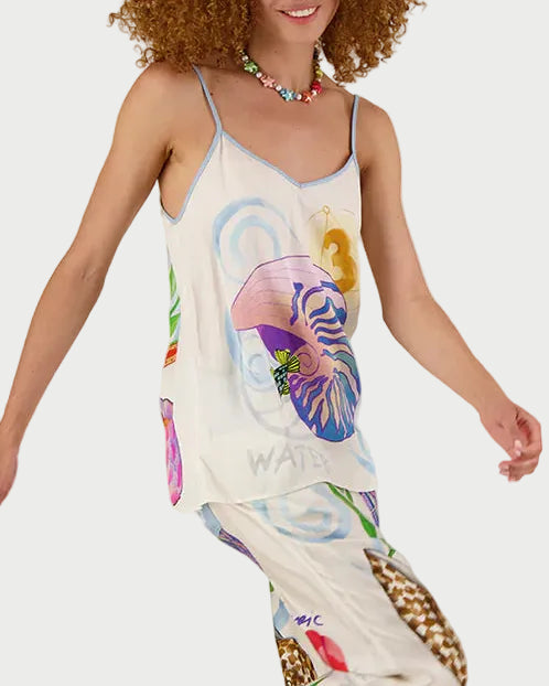 A smiling woman with curly hair wears the ME369 Angelique Camisole, a colorful, patterned top with seashell and ocean-inspired designs, paired with a beaded necklace. She poses in motion against a white background.
