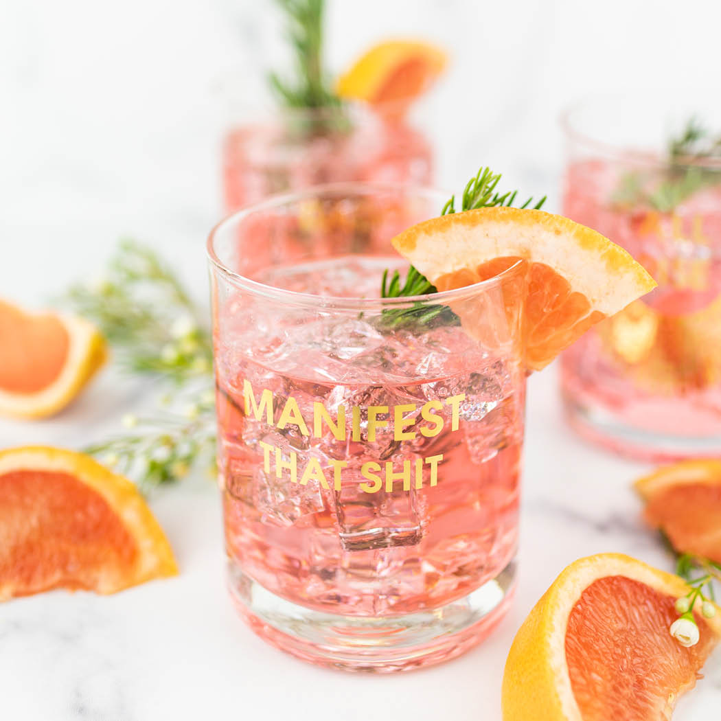 A Chez Gagné More Rocks Glass holds a pink cocktail, garnished with grapefruit and rosemary. The glass features the gold foil imprint "Manifest That Shit." More cocktails and grapefruit slices are seen in the background.