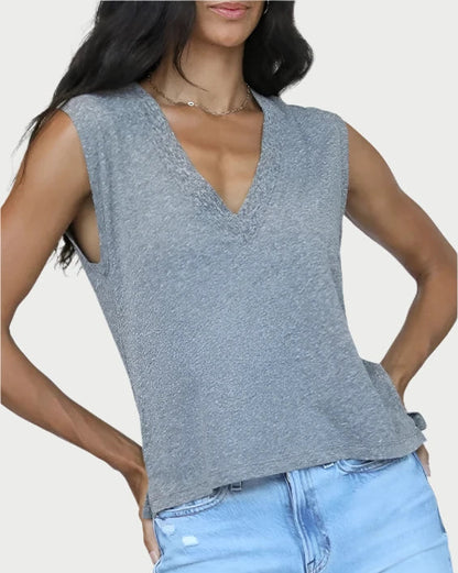 A woman in a gray Margot V Neck Sleeveless Tee by perfectwhitetee, paired with light blue jeans. Her long dark hair cascades down as she stands confidently with one hand in her pocket against an off-white background, effortlessly showcasing a flattering basic style.