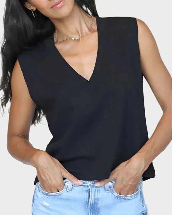 A person in a black Margot V Neck Sleeveless Tee by perfectwhitetee and light blue jeans, with hands casually in pockets. Their long, wavy dark hair complements the top, and they accessorize with a delicate necklace and hoop earrings, standing against a plain background.