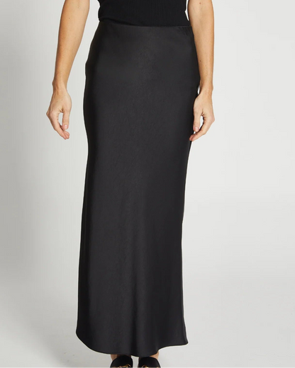 A person in a sleeveless black top and the Saint Art Charmeuse Maxi Skirt in Black by Saint Art NY, a long satin skirt, stands against a plain white background.