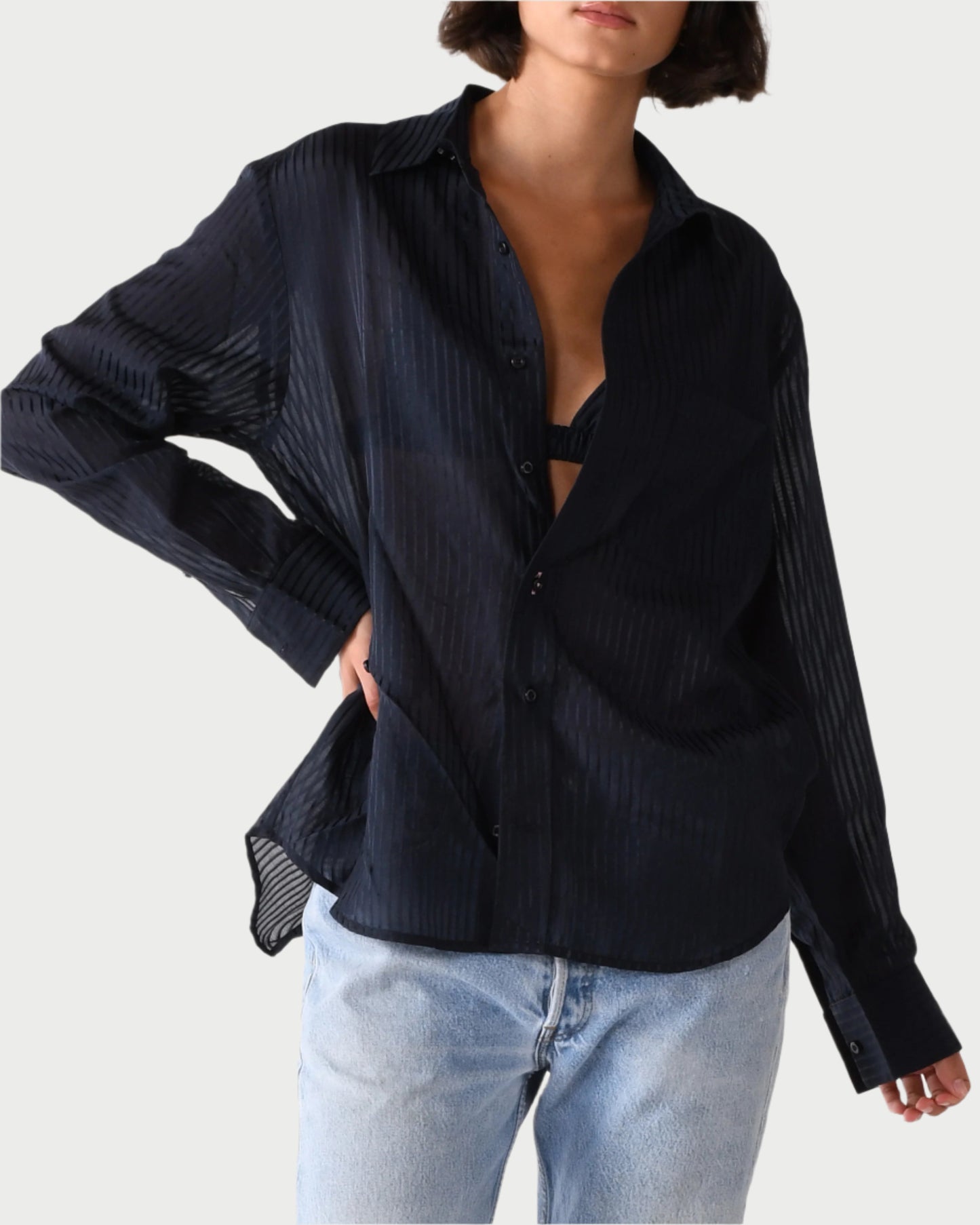 A person wears WEAR CISSA’s The Men's Shirt, a loose, semi-sheer black button-down with a subtle striped pattern over a black bra and light blue jeans. Their left hand is in their pocket against a plain white background.