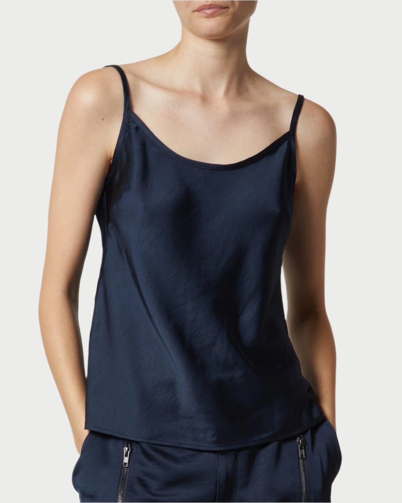 A person in a navy Charmeuse Camisole by Saint Art NY with matching trousers featuring zippered pockets stands against a plain background, hands in pockets, adding versatile color to their look. Their face is partially visible.
