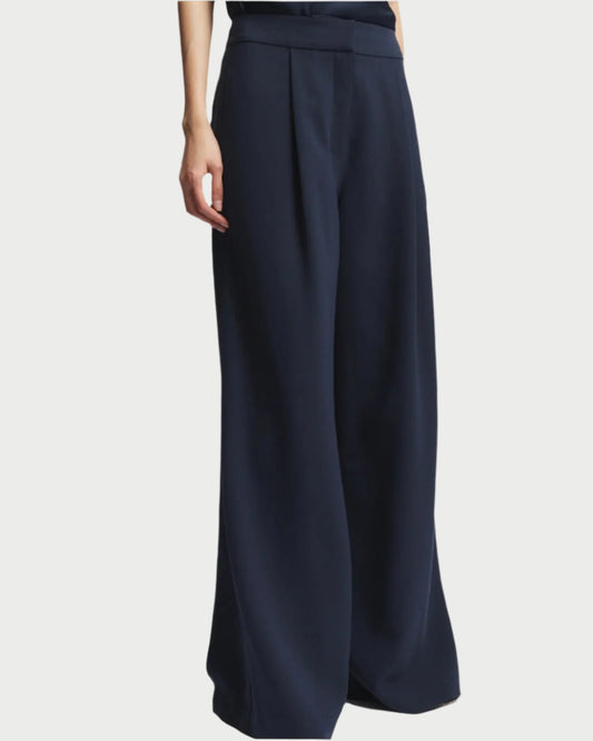 A person in the stylish Neve Trouser by Saint Art NY, high-waisted with a wide-leg and pleated front. Crafted from wrinkle-resistant navy crepe, the trousers flow elegantly. The hand rests casually against a plain white backdrop.