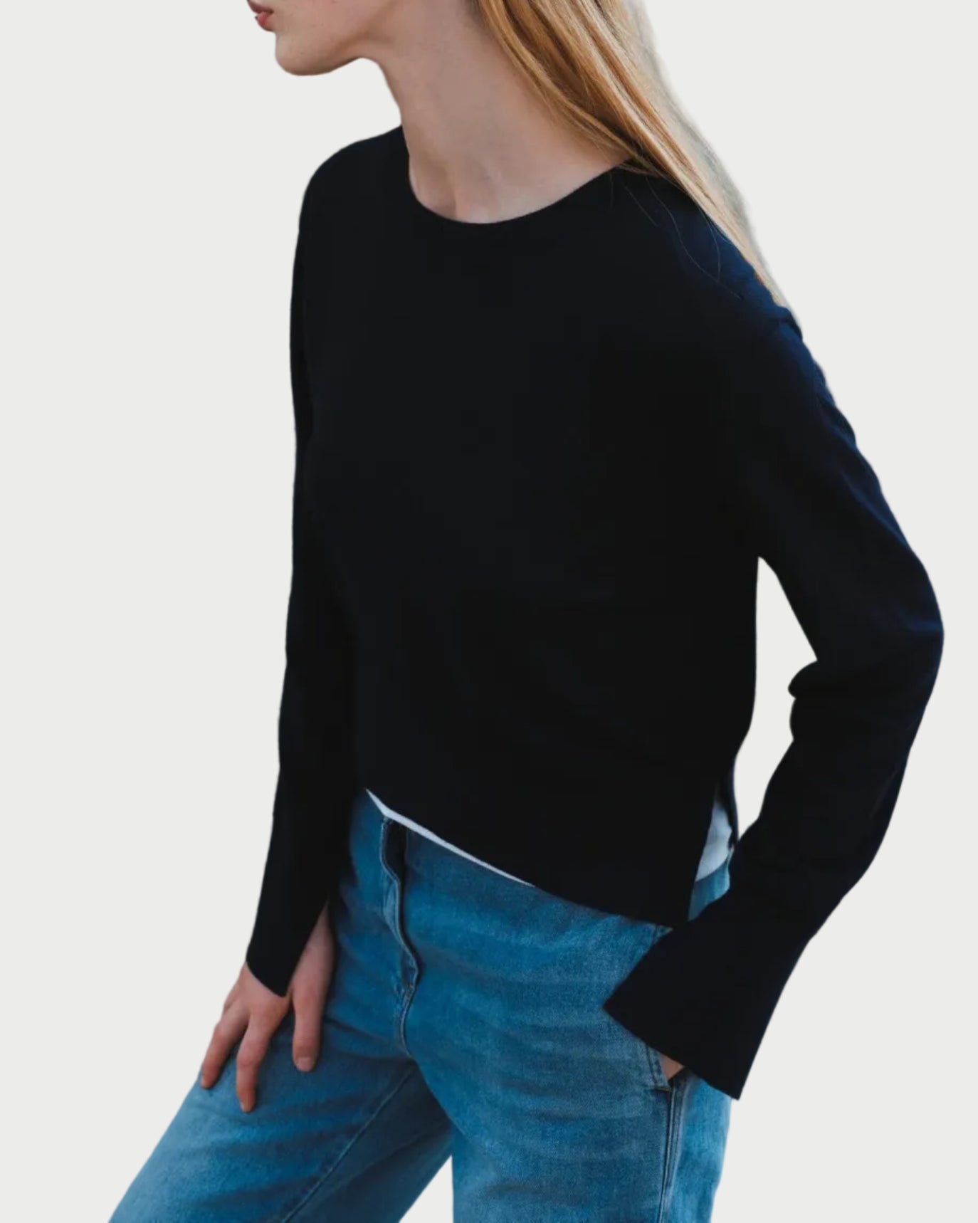 A person with long blond hair wears the navy Saint Art Elodie Sweater by Saint Art NY and blue jeans, posing with one hand in a pocket against a white background.
