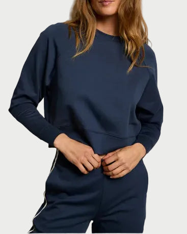 A woman wears a navy Perfectwhitetee Nell Travel Crewneck Sweatshirt from perfectwhitetee, paired with matching pants with white side stripes. She stands against a plain background, adjusting the sweatshirt at her waist with both hands.