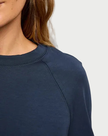 Cropped image of a person with long brown hair wearing the perfectwhitetee Nell Travel Crewneck Sweatshirt in dark blue heavyweight fleece, showing part of the face, neck, and shoulder against a plain light background.