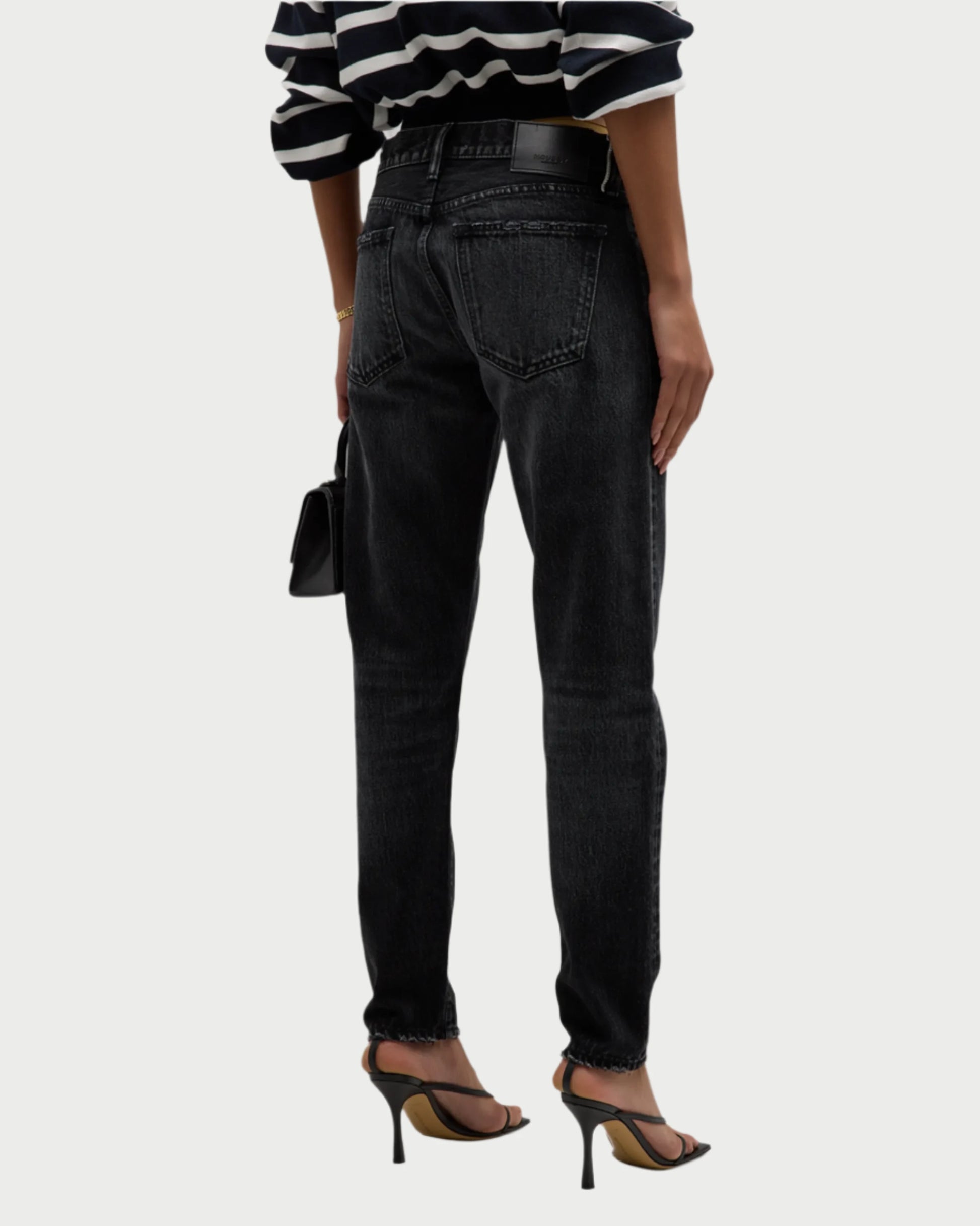 A person dressed in a black and white striped sweater and Vellflower Jeans by Moussy, which feature a high-rise waist and straight-leg fit, holds a small black purse. They are standing in black high-heeled sandals with their back turned.