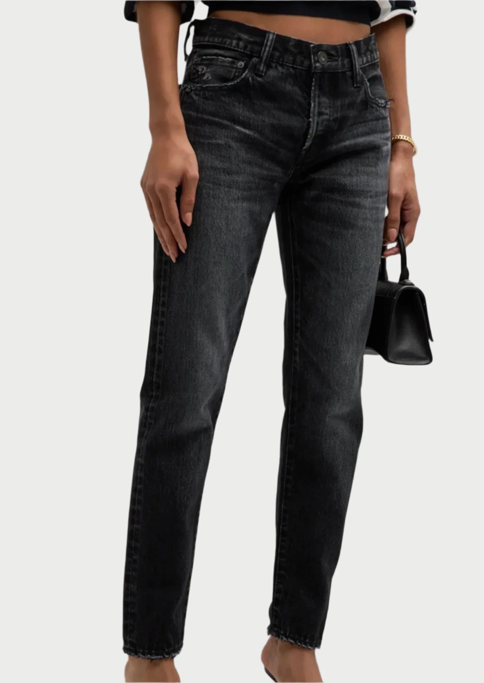 A person wearing Moussy's Vellflower Jeans, which are straight-leg black denim jeans with a high-rise waist and a faded design, paired with a black and white striped top. They are holding a small black handbag in front of a plain, light-colored background.