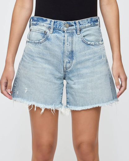 A person wears Moussy’s Nolita Denim Shorts—high-waisted, light blue cotton with frayed hems, slight distressing, front pockets, and button closure—paired with a black top for the perfect summer look.