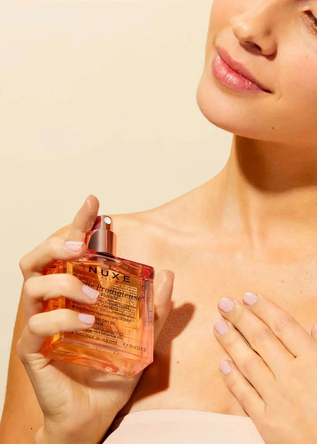 A person with bare shoulders and minimal makeup applies NUXE Nuxe Florale Dry Body Oil, a multi-purpose dry oil, on their collarbone. The floral scent enhances the moment as they smile slightly against a neutral background. The bottle contains a golden liquid and features a spray nozzle.