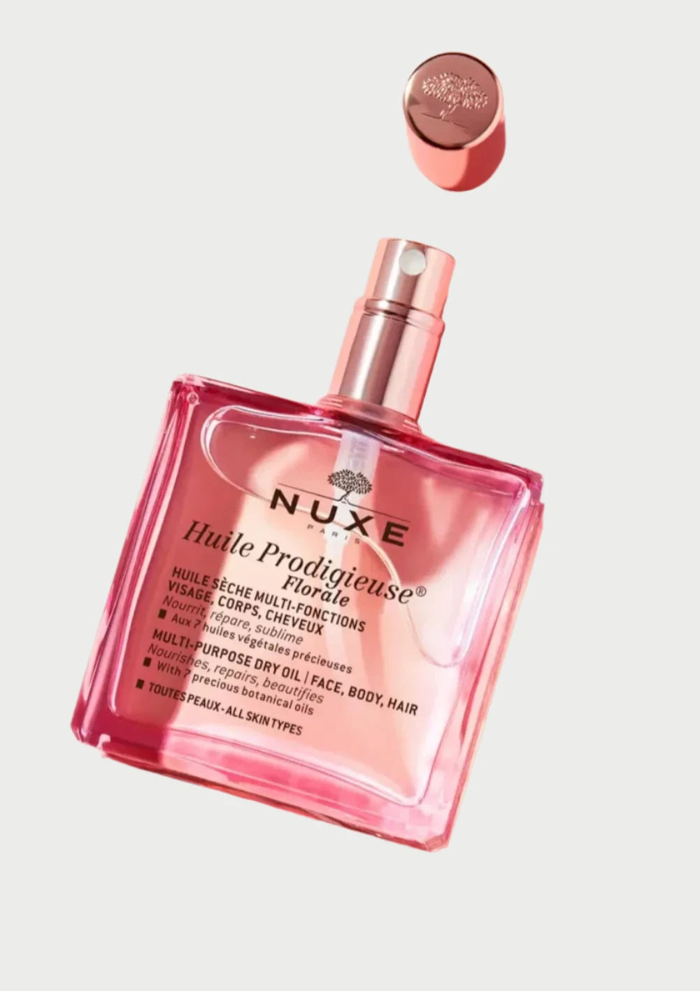 A bottle of NUXE Nuxe Florale Dry Body Oil, with its cap beside it, rests on a pink background. The rectangular glass bottle contains pink-tinted oil with English and French text detailing its uses for face, body, and hair. Enjoy the floral scent and nourishing botanical oils it offers.