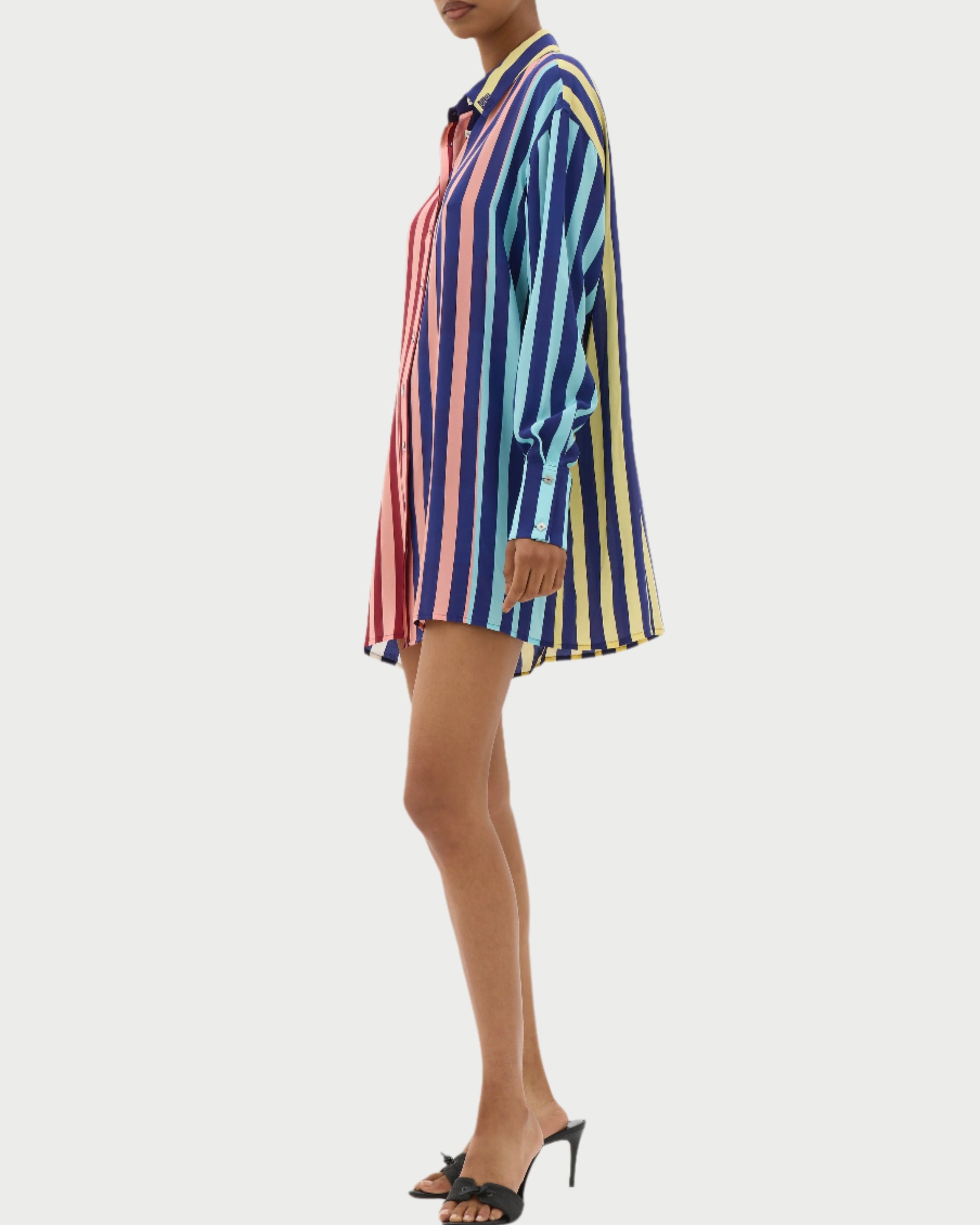 Against a plain background, a person wears the MUMA Striped Oversized Shirt as a vibrant dress featuring blue, yellow, and pink vertical stripes. Its long sleeves and pointed collar pair well with black high-heeled sandals for a chic take on the classic boyfriend blouse style.