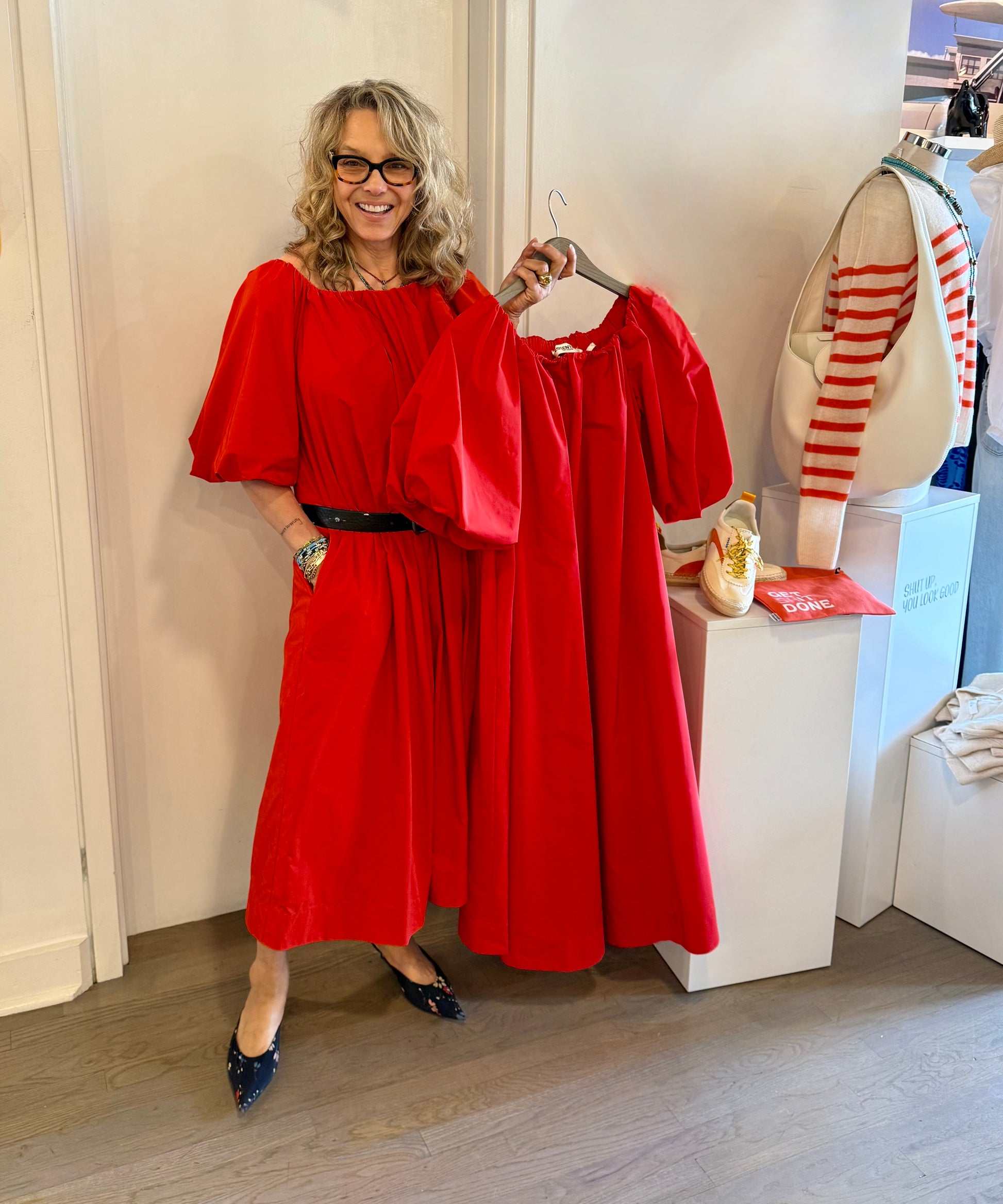 A smiling woman with wavy hair wears an Essentiel Antwerp Off Shoulder Midi Dress in red with black shoes indoors, holding a matching recycled polyester dress beside a white table displaying shoes, a bag, and a red-and-white striped sweater.