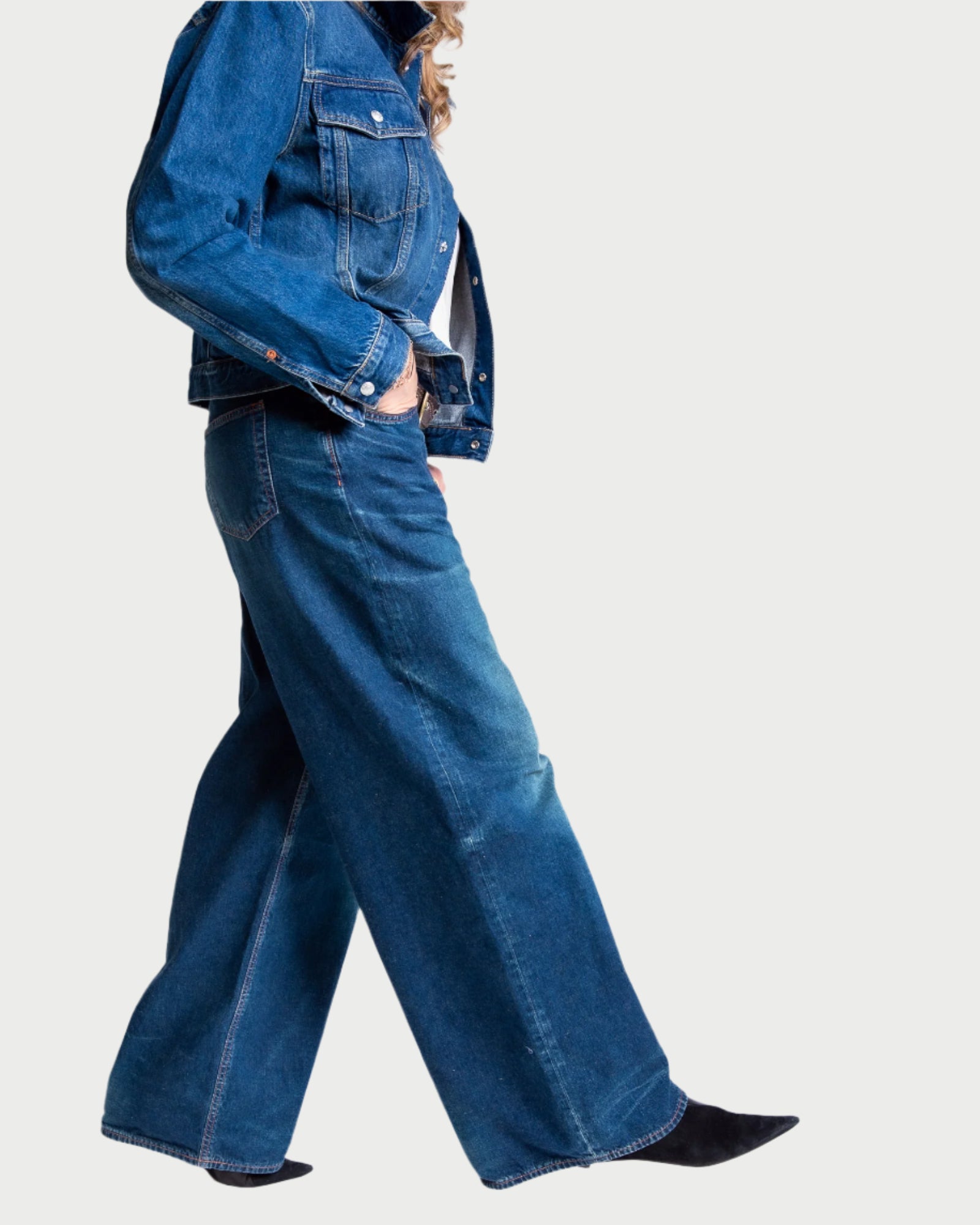 Against a white background, a person in a blue 100% cotton denim jacket and Moussy's Emporia Super Wide Leg Jeans embodies classic denim-on-denim style with one hand in their pocket, complemented by black heeled shoes.