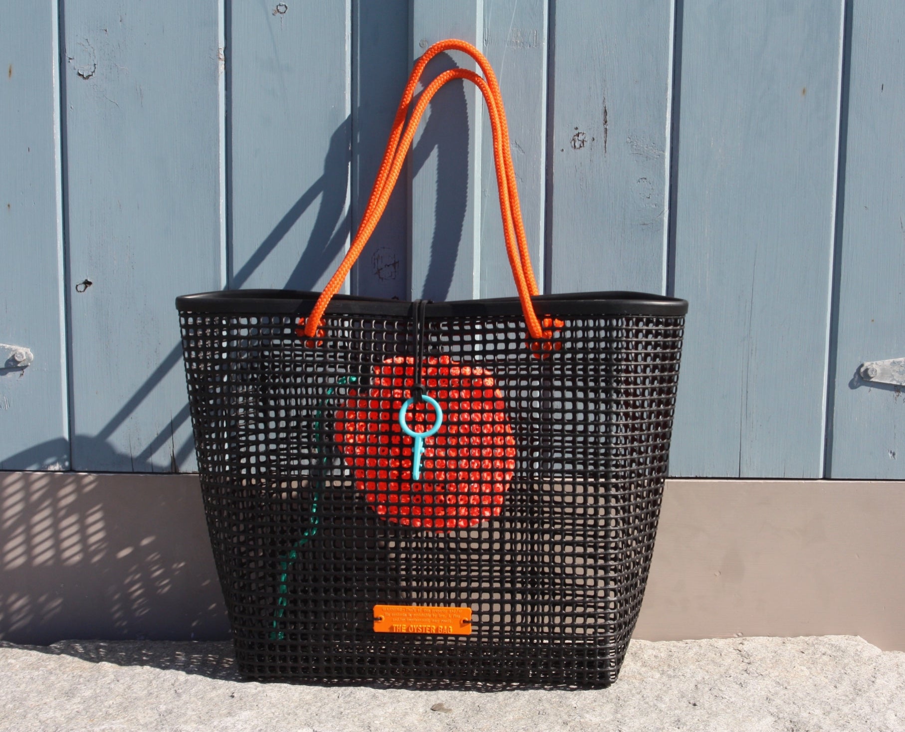 The Market Bag by THE OYSTER BAG—a black handmade mesh bag with bright orange handles and an orange item inside—leans against a light blue wooden wall on stone, perfect as your next beach tote.