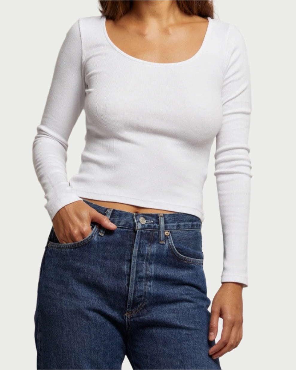 A person wearing the Anya Ribbed Long Sleeve Tee by perfectwhitetee with high-waisted blue jeans stands against a plain background, one hand in a pocket, highlighting the chic versatility of this ensemble.