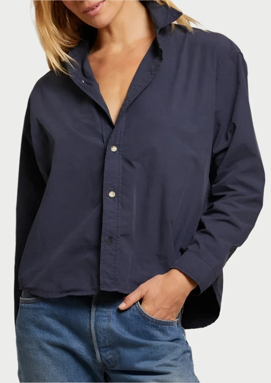 A woman with straight blonde hair is standing against a plain backdrop. She is wearing an effortlessly stylish James Button Down Shirt in navy blue from perfectwhitetee, with one hand in her pocket and blue jeans. She is looking at the camera and smiling slightly.
