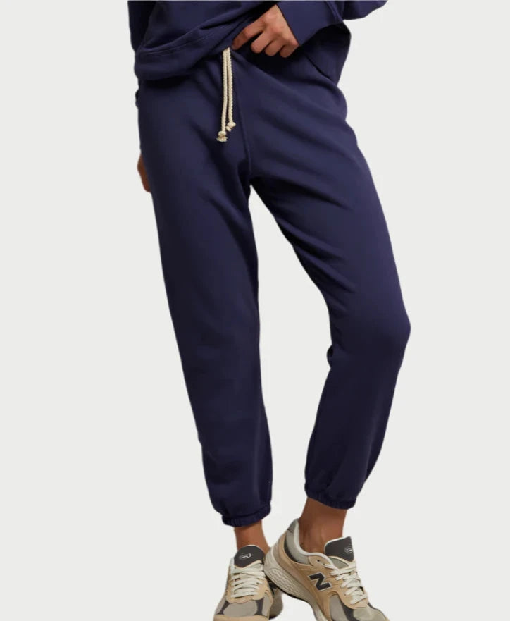 Dressed in the Johnny Sweatpant in Dusk Blue by perfectwhitetee, made from French terry cotton, an individual confidently poses against a plain backdrop. The sweatpants feature a drawstring waist and cuffed ankles, exuding a cool California vibe when paired with white sneakers and a casual white tee.