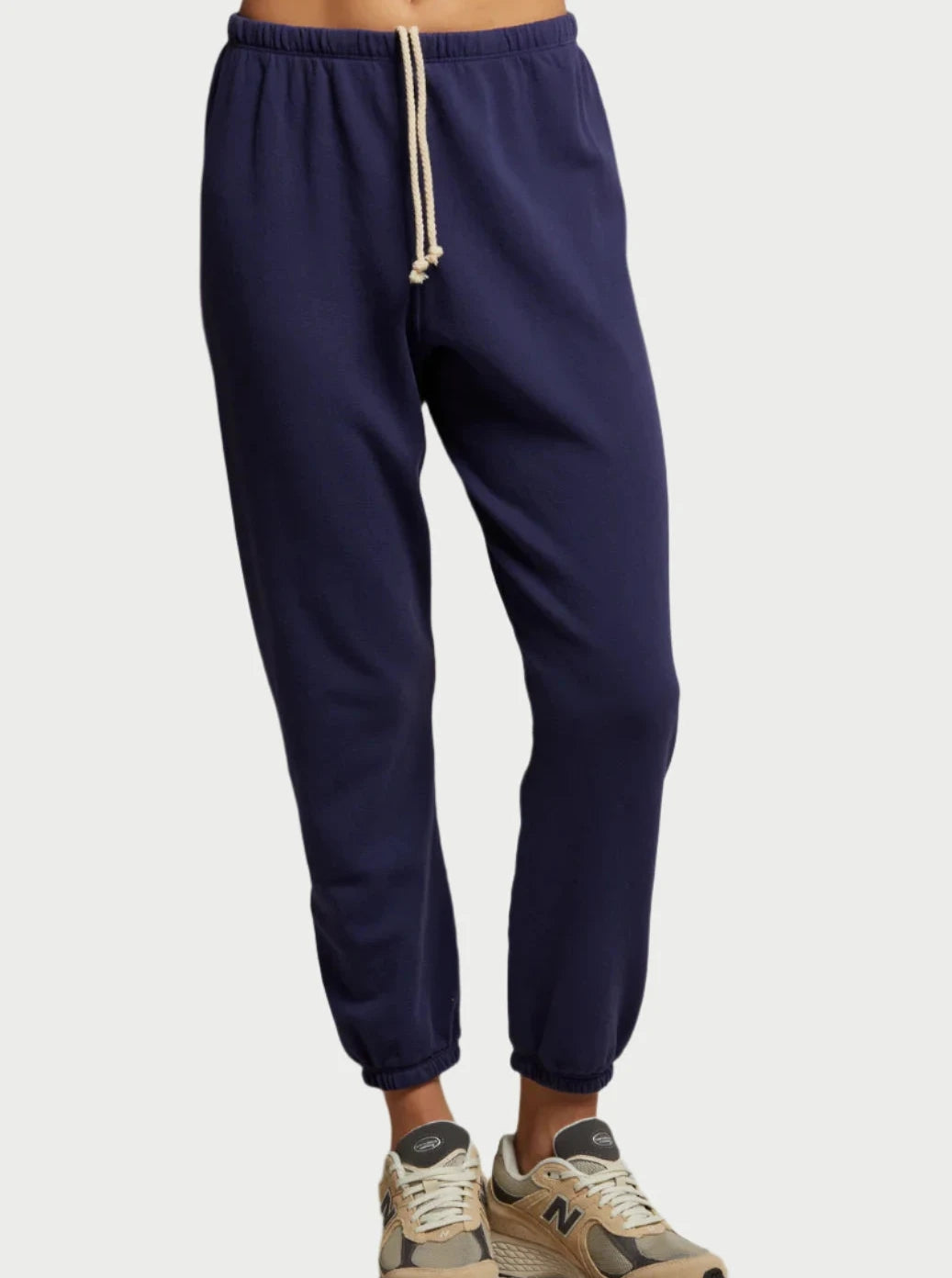 A person wearing the Johnny Sweatpant in Dusk Blue by perfectwhitetee, crafted from French terry cotton and showcasing an elastic waistband with a beige drawstring, paired with beige and gray sneakers. The image focuses on the lower half of their body against a light background, evoking a laid-back California vibe.