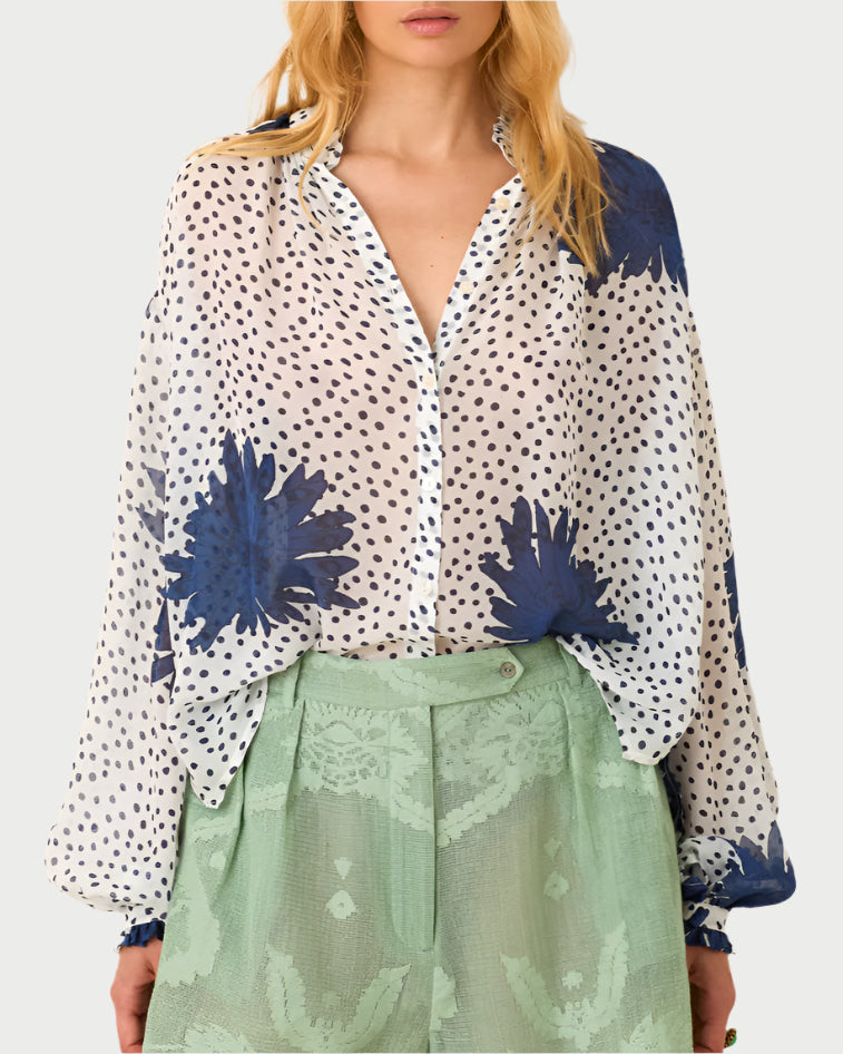 A person wears the Alix of Bohemia Poet Dahlia Blouse, a sheer, vintage-inspired top with blue floral patterns and polka dots, featuring a ruffled collar. It's tucked into high-waisted translucent green lace pants. The individual has long light hair and stands against a plain backdrop.