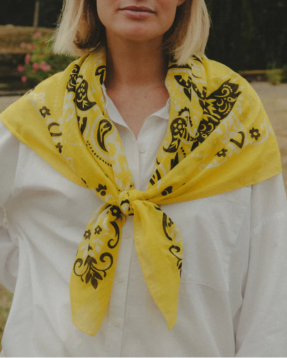 A woman wears a white button-up shirt and the Maison Lecomte Flament Bandana Scarf by Maison Lecomte Flament, tied around her shoulders as a lightweight accessory. She stands outdoors, her face partially visible from the nose down.