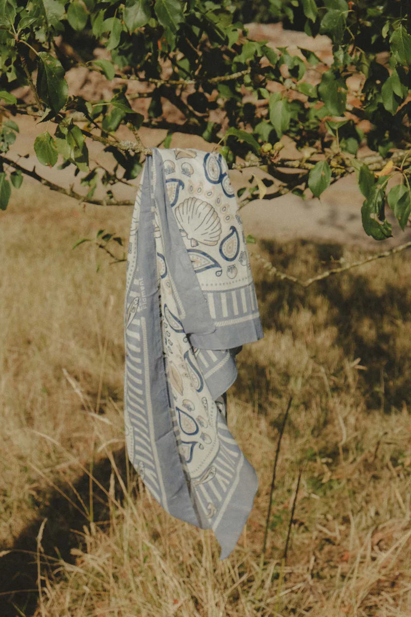 The Maison Lecomte Flament Bretagne Scarf by Maison Lecomte Flament, in blue and beige with a maritime print, is draped over a low tree branch in a grassy outdoor setting, sunlight casting soft shadows on the ground.