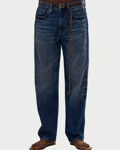 A person wearing R13 Boyfriend Jean by R13—relaxed fit, mid-rise blue jeans with faded details—paired with a brown belt and brown flip-flops, standing against a plain white background.