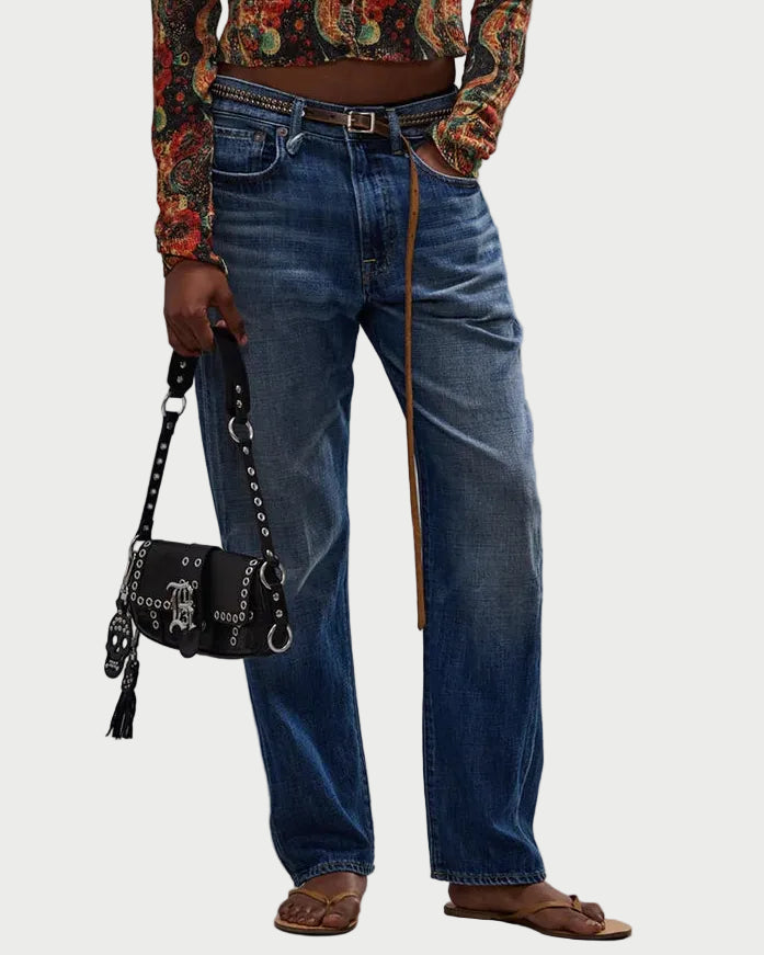 An individual wearing R13 Boyfriend Jean in blue, a patterned long-sleeve shirt, brown sandals, and a long brown belt. They carry a black studded handbag with a tassel.