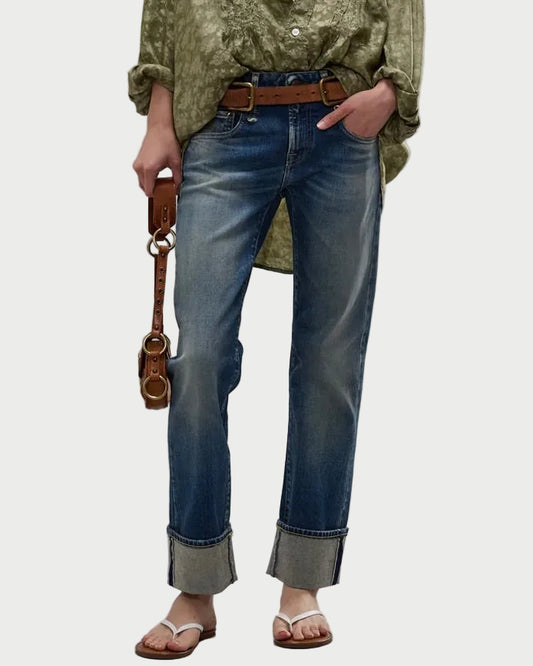 A person wearing blue straight-leg jeans with a faded wash and rolled cuffs, styled with a brown belt, olive green top, and brown sandals, holding a small brown leather bag against a light gray studio background.