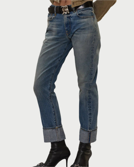 A person wears R13 Cuffed Romeo Jeans in Merrin Blue with a relaxed, worn-in look, styled with a black belt and black heeled boots, standing against a white background.