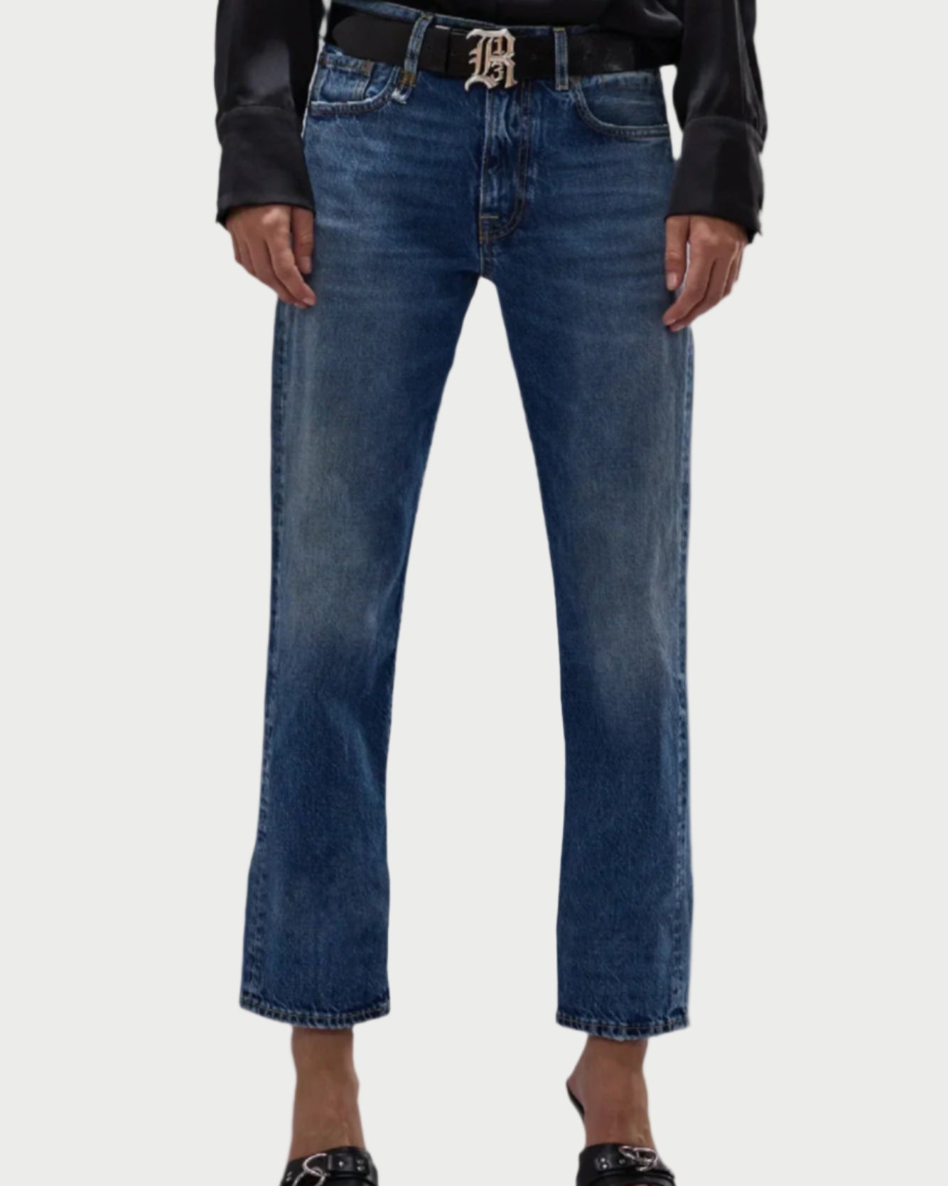 A person in an R13 Romeo Jean by R13, paired with a 100% cotton black shirt and black open-toed sandals, accessorized with a black belt featuring a decorative buckle. The plain light-colored background accentuates the elegance of this Made in Italy ensemble.